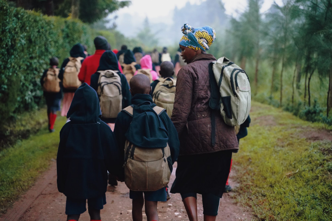 An adult and two children walk down a dirt road with back packs on behind a larger group of people