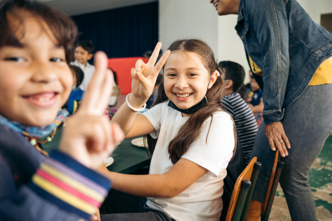 Two children holding up peace signs and smiling in a classroom setting